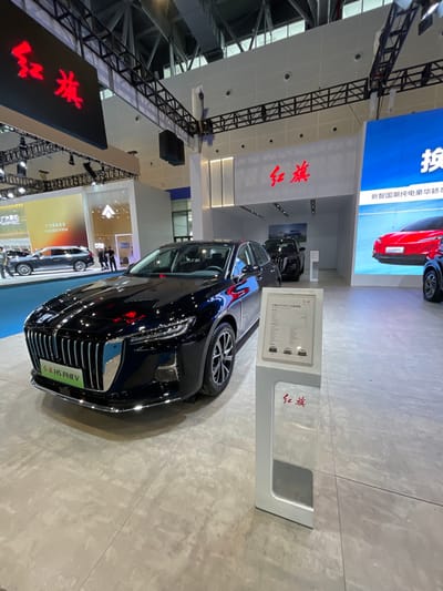 The image shows a black Geely H5 PHEV car on display in a large, brightly lit showroom. The car is positioned in the center of the image, facing the viewer, with its front grill and headlights prominently visible. The car is parked on a grey tiled floor. To the right of the car is a white stand with a sign that reads "Geely" in red Chinese characters. The stand also displays a technical specification sheet for the car. Behind the car is a white wall with a sign that reads "Geely" in red Chinese characters and the words "New Energy and Fuel Vehicles" written in English. The showroom is sparsely decorated and has a high ceiling with a large network of metal girders. There are other cars visible in the background, including a red car and a dark SUV. The image was taken in Hangzhou, China, at the coordinates 30.272 latitude and 120.416 longitude. The scene is empty of people.