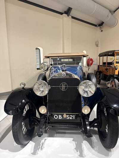 The image depicts a vintage blue and black car, possibly on display in a museum. The license plate reads "OB 521." The car is positioned in front of a neutral-colored wall. An overhead ventilation system is visible near the ceiling. The location appears to be indoors, likely within the Stellenbosch Local Municipality, South Africa, based on the car's presence in a museum environment.