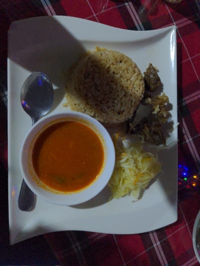 The image displays a meal served on a white, rectangular plate with gently curved edges. The setting appears to be a dimly lit indoor environment, possibly a restaurant or home, characterized by a red and white checkered tablecloth visible in the background and foreground.
The plate holds several food items:
- A generous mound of light brown, spiced rice, possibly pilau, positioned on the left side of the plate.
- To the upper right of the rice, there are dark, cooked pieces, likely meat or a rich stew.
- Adjacent to the meat, a portion of finely shredded, light-colored vegetables, possibly cabbage or a similar slaw.
- A small white bowl, placed centrally at the bottom of the plate, contains a vibrant orange-red soup or sauce. This sauce appears to have faint green specks, possibly herbs or spices.
- A silver spoon rests beside the bowl of soup, positioned on the plate.
The lighting is soft and appears to come from the top-left, casting noticeable shadows from the food items onto the plate. In the upper right corner of the background, out-of-focus colorful lights (bokeh) are visible, suggesting decorative lighting often found in evening settings. There is no visible text in the image. Given the location context of Bagamoyo, Tanzania, the meal likely represents local Tanzanian cuisine.