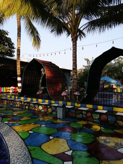 The image depicts an outdoor scene at a water park or recreational area in Kebonsari, Indonesia. In the foreground, a vibrantly colored, mosaic-like pavement is visible, featuring large, irregularly shaped sections of green, yellow, blue, and red. A curved stone border separates the colored pavement from a blue pool of water.
Dominating the mid-ground are two large, arched, dark structures, possibly gazebos or shaded seating areas, with curved roofs resembling dark brown tiles. One structure is more fully visible on the left, showcasing a dark, open interior with a visible grill or fence. The other arched structure is on the right, partially obscuring a swimming pool.
Several palm trees are present, with their fronds extending across the upper portion of the image, casting some shade. String lights with small, colorful bulbs are suspended between poles or structures in the background, suggesting an evening or decorative lighting setup.
Further in the background, there are structures with colorful fences and what appear to be decorative mushroom-shaped elements. A blue umbrella and a sign indicating "TOILET" are also visible, suggesting the presence of amenities. The sky is clear and pale blue, indicating daytime.
The scene is set in an outdoor environment, likely a water park or a similar leisure facility. The overall impression is one of a colorful and playful atmosphere. There are no visible people actively engaged in any actions. The image captures a static view of the environment.