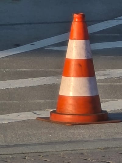 A solitary orange and white traffic cone stands on a paved surface. The cone has three orange sections and two white reflective bands. The base of the cone is black and circular. The cone is positioned on a grey, asphalt-like surface with faint white stripes suggesting a road or pedestrian crossing. The background consists of more grey paving with additional parallel white stripes. The lighting suggests it is daytime, casting a shadow from the cone. No people or other objects are visible in the immediate vicinity. The scene is likely in the city of Flörsheim, Germany, given the context.
