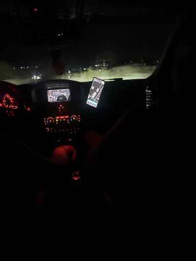 The image depicts the interior of a car at night, possibly while driving. A smartphone displaying a map or navigation app is mounted on the dashboard. The car's dashboard displays illuminated controls and gauges. The exterior of the car is dimly visible through the windshield, showing city lights. The location on the navigation app appears to be close to Petarch, Bulgaria. The driver's hand is visible on the steering wheel.