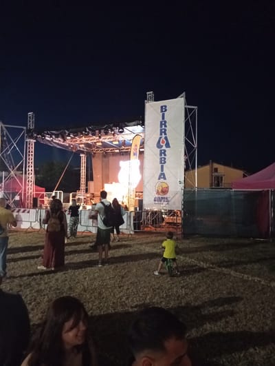 The image depicts an outdoor event at night, likely a festival, in the city of Arbia, Italy. A stage is set up with lighting rigs and a large screen displaying dynamic visuals, possibly a performance or concert. A prominent white banner with blue lettering reads "BIRRABBIA," indicating a beer-related event or festival. Below this text, there is a logo with a beer mug and the text "AVIS Tavolne eu suada." Several people are visible in the foreground and mid-ground, some standing and observing the stage, while others are moving about. A young child on a balance bike is present in the mid-right of the frame. Tents and temporary fencing are set up around the event area, and the ground is covered in grass. The overall atmosphere suggests a lively community gathering.