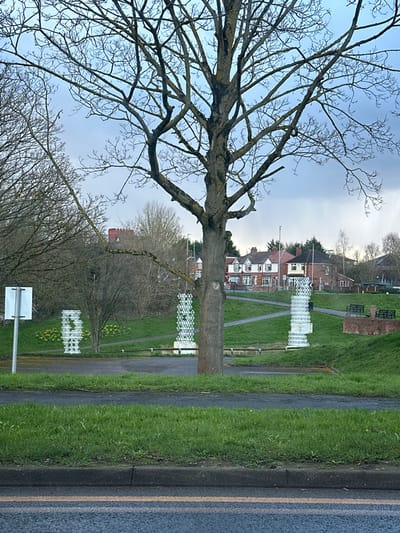 The image depicts an urban park scene in Winsford, United Kingdom, observed from across a roadway under an overcast sky. In the foreground, an asphalt road with a double yellow line is visible, bordered by a strip of mown green grass. A large, deciduous tree, bare of leaves, stands centrally in the mid-ground.

Beyond the tree, three white, open-lattice, columnar sculptures of varying heights are positioned on a green, upward-sloping lawn. A cluster of yellow daffodils is present near the leftmost sculpture. A winding paved footpath traverses the grassy area. To the left, a white square sign on a metal pole is visible. Further to the right and slightly elevated within the park, two dark park benches and a circular brick planter are present.

In the background, a row of residential brick houses with tiled roofs is visible, interspersed with streetlights. A prominent red-brick building featuring a castellated roofline stands in the distant left. The scene is devoid of individuals or active events.