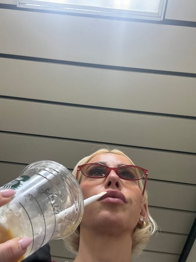 The photograph shows a woman holding a Starbucks cup with "EMILY" written on it. She is drinking from the cup through a straw and wearing red eyeglasses. She has blonde hair and is wearing earrings. The background appears to be a wall with vertical paneling. The location in Ferno, Italy is not determinable from the photograph.