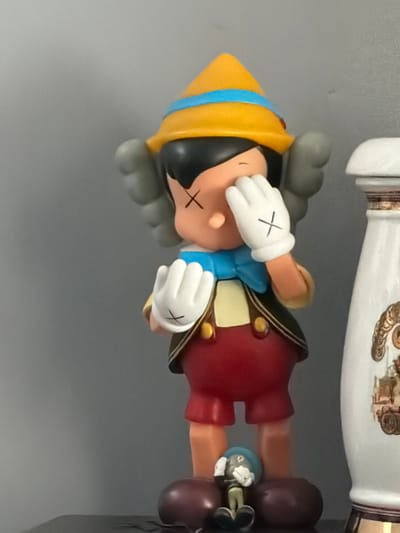 A Pinocchio figurine is shown, lying horizontally with its head to the left. Its eyes are covered with white gloves marked with black 'X's. The figurine wears a yellow hat with a blue band, a blue bow, a brown and beige jacket, and red pants. The figure's face also bears a black 'X' mark. A decorative white and gold vase is visible in the background, partially out of focus. The figurine is resting on a gray surface.