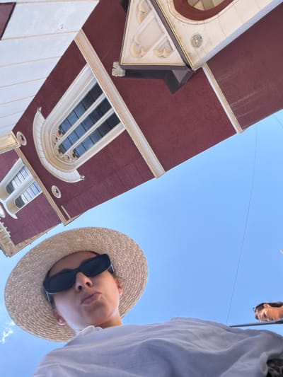 A low-angle, wide shot captures a woman wearing a straw hat and sunglasses, looking directly at the camera with her lips pursed. She is dressed in a white top. Behind her, a portion of a red building with white architectural details and windows is visible, tilted and appearing to hang from the sky. The building's design suggests it could be part of a historical or religious structure, potentially resembling a church or a grand old building. The background is a clear, bright blue sky. To the right, another person's head and shoulders are visible, looking down and to the side, also against the blue sky. The scene is outdoors, likely in a city, and the perspective suggests the photo was taken from a low vantage point looking upwards. Based on the architectural style and the general atmosphere, it is plausible that this scene is located in Sarajevo, Bosnia and Herzegovina, a city known for its rich history and blend of architectural influences.