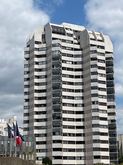 The image shows the exterior of a modern, tall, multi-story building in Créteil, France. The building is constructed with a repeating pattern of light-colored panels and darker bands that run diagonally across its facade. Numerous windows are visible on each level, some of which appear to be balconies or recessed areas. The top of the building features an angular, stepped design with several protruding sections. In the foreground, several flags are visible on poles, including a red, white, and blue flag. The sky above is partly cloudy, with patches of blue visible. The scene appears to be an urban environment, with other buildings partially visible in the background. There are no people clearly visible in the image.