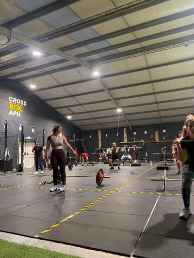 Inside a gym, individuals exercise in a structured setting. Some participants are engaged in weightlifting near squat racks marked by yellow lines. A woman in sportswear appears to be demonstrating or warming up, with her arm extended. The gym has a high ceiling with visible metal beams and bright lighting, contrasting against the dark wall with "CROSS BOX APH" printed on it. The floor is covered in dark rubber mats.