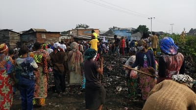 The image displays a daytime scene in an informal settlement within Gisenyi, Rwanda. A dense gathering of approximately 50 to 70 individuals, predominantly adults, is observed across an expansive dark debris field, consistent with the aftermath of a fire or significant waste accumulation. Several individuals in the foreground and midground are seen interacting with the debris, some using wooden poles or sticks; one person in the mid-right wears a white face mask. In the background, numerous densely packed, informally constructed residential structures, fabricated from wood and corrugated metal, form the settlement. One building exhibits blue paint. Overhead power lines and streetlights are visible in the upper right quadrant. Sparse green vegetation borders the scene on the far right. The sky appears overcast or hazy.