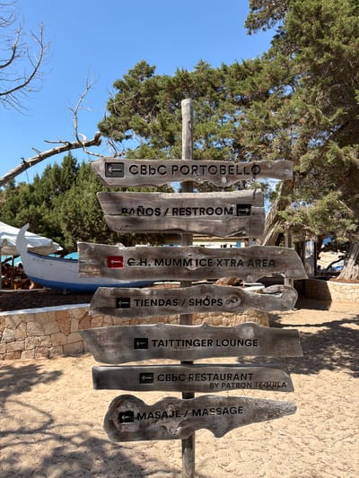 This media file features a wooden signpost with multiple signs indicating directions to various establishments. The signs are made of weathered wood with carved text and directional arrows. From left to right, the signs read: "CBBC PORTOBELLO" with a left arrow, "PAÑOS / RESTROOM" with a left arrow and a small icon, "CH. MUMM ICE XTRA AREA" with a red tag icon, "TIENDAS / SHOPS" with a left arrow, "TAITTINGER LOUNGE" with a left arrow, "CBBC RESTAURANT BY PATRON TEQUILA" with a left arrow, and "MASAJE / MASSAGE" with a left arrow and a small icon. The signpost is set against a backdrop of a blue sky, lush green trees, and a textured wall made of stone and stucco. The ground appears to be sandy. A boat, partially covered, is visible behind the signpost. The lighting suggests it is daytime, with shadows cast on the wall. The overall scene evokes a relaxed, outdoor atmosphere, likely at a beach resort or a similar leisure destination in a sunny climate, consistent with the provided location context of Sant Josep de sa Talaia, Spain. There are no people visible in the image.