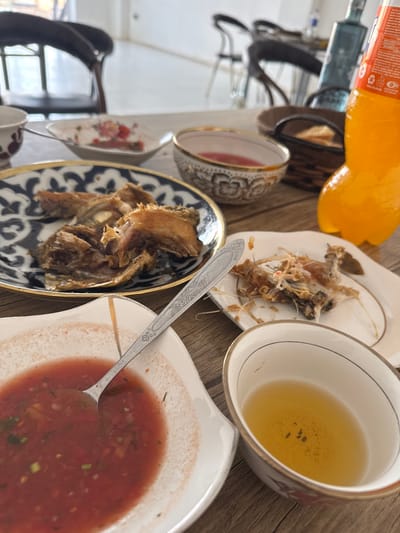 The image depicts a dining table laden with dishes after a meal in Kasan, Uzbekistan. The primary subjects are various food remnants and containers arranged on a light-colored wooden table. A prominent round plate with a dark blue and white Uzbek-style pattern holds several pieces of partially consumed fried fish. Adjacent to it, a small white rectangular plate with gold trim contains fish bones and skin, indicating completed consumption. A rectangular white bowl, also with gold trim, contains a red, chunky sauce with green herbs, and a silver spoon with an ornate handle rests within it. To the right, a smaller, ornate bowl holds a clear, light amber liquid, likely tea, with a few dark particulates at the bottom. Behind these, a small, patterned ceramic bowl contains a pinkish liquid. A 1.5-liter plastic bottle of Fanta orange soda, approximately half full, stands upright to the right of the dishes. A small, woven brown basket is visible behind the Fanta bottle. In the blurred background, two dark wooden chairs with light seats are positioned around a larger, white table, and a tall blue glass bottle is partially visible. The overall environment suggests an indoor dining area with natural light. No individuals are visible.