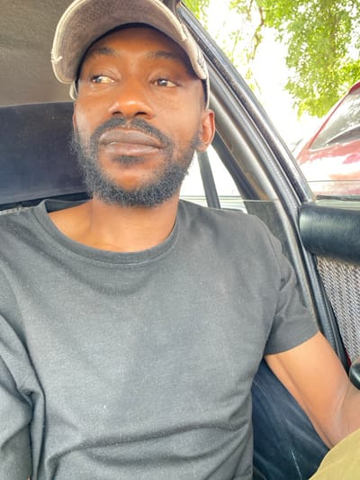 A male individual is depicted seated within a vehicle, located in Ungwan Liman, Nigeria. He is observed from the chest up, wearing a light-colored baseball cap and a plain dark short-sleeved t-shirt. The individual has dark skin, a neatly trimmed beard, and a mustache. His head is turned slightly to the left, and his gaze is directed towards the upper left. Visible vehicle interior components include a dark door frame, a black armrest, and a gray patterned fabric panel on the inner door. Through the open window or windshield, green foliage and a portion of a red vehicle are discernible in the background, indicating an outdoor, daylight environment.