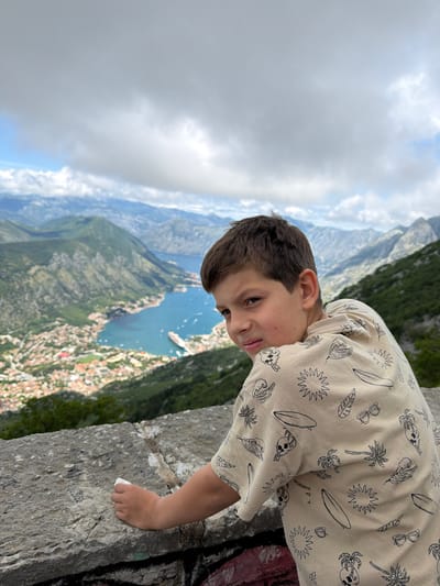 A young boy with dark brown hair and dark eyes is captured leaning over a weathered stone wall, looking downwards and sideways into a vast landscape. The image is rotated, showing the boy's head tilted towards the camera with a playful expression, including a slight smirk and his tongue peeking out. He wears a light-colored, long-sleeved hooded shirt adorned with black outline patterns of suns, palm trees, surfboards, sunglasses, and skulls. His right hand rests casually on the stone wall.
Beyond the boy and the foreground wall, a stunning panoramic view unfolds. A large, deep blue body of water, consistent with a bay or fjord, is visible, reflecting the partially cloudy sky. Along the shores of the water, a town with numerous buildings, some featuring prominent red roofs, is nestled, extending up the surrounding green, steep mountainsides. Several small boats dot the water. The sky shows a mix of white clouds and brighter areas where sunlight is breaking through, suggesting it is daytime. The scene strongly depicts the natural beauty of a coastal mountainous region, specifically aligning with the panoramic views found in Špiljari, Montenegro, overlooking the Bay of Kotor. The boy's pose and expression convey a sense of curiosity and engagement with the impressive scenery below.