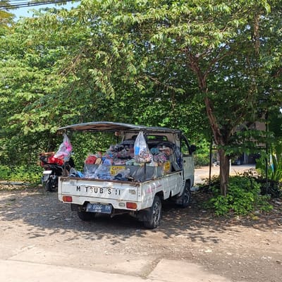 In Kebondalem, Indonesia, a white Mitsubishi pickup truck is parked on a dirt and gravel lot, likely a roadside or open area. The truck bed is covered by a canopy and filled with merchandise, primarily bags of footwear such as sandals and flip-flops, and some pink plastic shoes hang from the canopy. A motorcycle is parked next to the truck. The scene is shaded by a large tree with dense green foliage. The ground is covered with dirt, gravel, and scattered leaves, and the background shows more greenery and a hint of a building. A license plate on the rear of the truck reads "H 8956 RM".