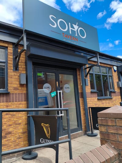 This is a medium-shot, eye-level photograph of the exterior of a pub called "Soho Tavern" located in West Bromwich, Sandwell, United Kingdom. The pub has a distinctive yellow brick facade. Above the entrance, a prominent light blue sign displays "SOHO TAVERN" in white lettering, with "WEST BROMWICH" below it.
The entrance to the pub features a dark-colored door with glass panels. A small green sign on the door reads "OPEN." Inside the glass, a white circular sign with "SOHO" is visible. To the left of the entrance, a window is partially obscured by blinds.
In front of the pub, a black barrier with chrome poles encloses a small outdoor seating area. A black banner displaying the Guinness logo and "GUINNESS" is positioned behind the barrier. The ground in front of the pub is paved with concrete.
The sky above is a vibrant blue with fluffy white clouds, suggesting it is daytime with pleasant weather. The overall impression is of a well-maintained and inviting local pub. There are no people visible in the image.
