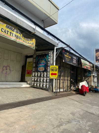 The image shows a street scene in Kecamatan Dayeuhkolot, Indonesia. The scene is a street corner with a shop on the left side. The shop is labeled "NAZAR PAINT CAT TEMBOK KILOAN". There are many cans of paint stacked on the wall of the shop. There is a sign above the door of the shop in Indonesian, which is not translatable in this context. There is a yellow sign on the right side of the shop that reads "NAZAR PAINT TOKO CAT". The sign has a red arrow pointing to the right. The street corner is concrete and there are several buildings and shops across the street. There is a motorbike with a white cover parked on the street. There is a power line above the shops. The sky is cloudy.
