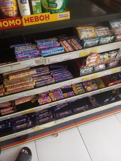The image displays an interior view of a convenience store aisle in Naung Bo Oe, Myanmar. The scene focuses on five visible shelves stocked with various consumer goods. The uppermost shelf features a yellow promotional sign for "Alfamart" with the text "HEBOH," alongside "Mie Sedaap Cup" instant noodles, a white bottled product, and a green pouch labeled "10X BERSIHKAN LEMAK."

The second shelf from the top is predominantly filled with numerous rolls of "Mentos" mints in blue, purple, and other wrappers, interspersed with "Kopiko" coffee candy. To the right, "Strepsils" lozenges are displayed in yellow and blue packaging. The third shelf contains stacks of "SilverQueen Chunky" chocolate bars, additional "Mentos" rolls, and various colorful wrapped candies. The fourth shelf continues with "SilverQueen Crispy" bars and rows of "Chupa Chups" lollipops/candies. The lowest visible shelf is stocked with multiple "Dairy Milk Cashew Nut" chocolate bars. Small white labels with pricing, indicated in Indonesian Rupiah (Rp), are affixed to the front edge of each shelf.

The floor is composed of white square tiles with gray grout lines, bordered by a red and black striped baseboard directly beneath the shelving. In the lower-left corner, a human left foot clad in a black sandal is partially visible. A small, light-colored cylindrical object rests on the tiled floor near the lower-right corner. Minor dust and debris are present along the edge of the lowest shelf.