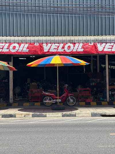 In Nathon, Thailand, a red motorcycle is parked on a sidewalk in front of a shop. The shop has a large red banner with white lettering that reads "VE LOIL VE". A colorful umbrella, with sections of red, yellow, and blue, shades the motorcycle and the sidewalk. The shop appears to be an auto repair or service center, with tools and equipment visible inside the open doorway. To the left of the main umbrella, another smaller, partially visible umbrella with red, green, and yellow sections is present. The sidewalk has black and yellow striped markings along the curb, and beyond that is a paved road. The building facade above the banner is constructed with corrugated metal. The scene is illuminated by bright sunlight.