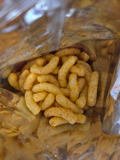 This image presents an internal, downward-angled view into an open snack bag, situated in Negorci, North Macedonia. The scene is dominated by a substantial quantity of light-yellow, puffed, corn-based snacks that are irregularly shaped, primarily curved cylindrical pieces, each exhibiting small, dark-brown seasoning specks. These snacks are densely packed at the bottom of the bag. The interior of the bag is lined with a highly reflective, crinkled metallic-foil material, likely a composite plastic or aluminum film, which reflects ambient light and creates varied bright and shadowed areas along its textured surface. The upper edges of the bag are visible, indicating its open state. No individuals or direct actions are depicted within the frame.