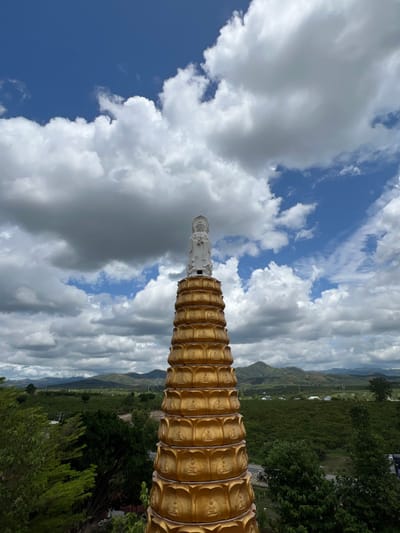 A tall, multi-tiered golden pagoda stands prominently in the foreground, topped with a white statue of a figure resembling Guan Yin, depicted with a halo and holding an object. The pagoda features multiple levels, each adorned with a lotus petal design and small, recessed Buddha figures. The scene is set against a backdrop of rolling green hills and mountains under a partly cloudy sky with patches of bright blue visible through large cumulus clouds. Lush green vegetation, including trees and what appears to be agricultural fields, covers the landscape. A road and scattered structures are discernible in the middle distance. This location is identified as the city of Đăk Mar, Vietnam.