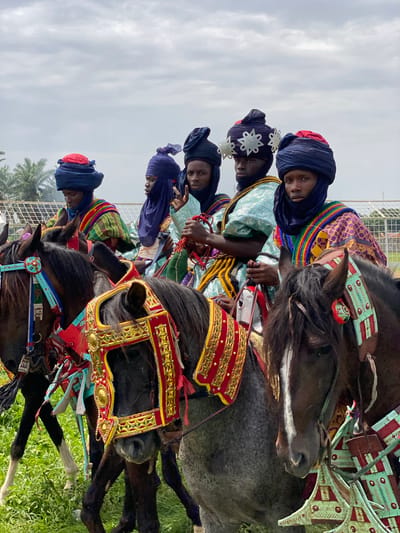 Five men wearing blue turbans and colorful, patterned robes are riding horses that are adorned with ornate saddles and decorative coverings. The men are lined up in a row, facing the same direction. The horses are mostly black with some grey. They are on a green grassy field. The sky is overcast and grey. In the background are some trees and a fence. The scene is a Durbar festival in Kafanchan A, Nigeria.