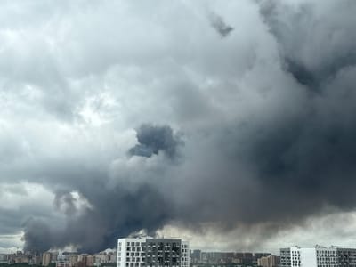 A wide shot captures a panoramic view of the Tomilino, Russia skyline under a heavily clouded, dark sky. The lower portion of the image shows a dense city landscape with numerous residential buildings of varying heights and architectural styles, interspersed with greenery. In the foreground, a modern, white apartment building with numerous windows stands out. Dominating the upper and central portions of the frame is a massive plume of dark, billowing smoke, originating from somewhere within the city and rising high into the atmosphere. The smoke appears thick and ominous, obscuring a large section of the sky and casting a somber mood over the scene. There are no visible people or specific actions depicted, other than the environmental event of the smoke.