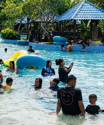 A group of people are enjoying a day at a public swimming pool, likely part of the Pantai Cermin Kiri tourism area in Indonesia. In the foreground, a man in a black t-shirt with a smiley face design sits in the water, facing away from the camera. Beside him, a young child in a black t-shirt also sits in the water. Several other individuals, including women and children, are visible in the pool, some swimming and others standing. In the midground, a woman is holding a smartphone, appearing to take a photo or video. There are inflatable pool toys, including a large yellow ring and a blue tube, in the water. In the background, there are trees and structures with blue roofs, characteristic of resort or park amenities. The water is clear blue, and the sunlight creates reflections on the surface.