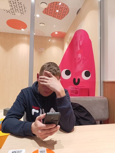 The image shows a young man sitting at a table in an establishment. He is wearing a dark blue hoodie with a "FILA" logo. The man has his right hand covering his eyes, and he is looking down at a smartphone held in his left hand. The background features a wall with a light wood grain pattern. Behind him, through a glass partition, is a large, pink, cartoonish character with two googly eyes and a U-shaped mouth. The ceiling of the establishment is decorated with circular patterns and abstract shapes. There are also some fluorescent lights visible on the ceiling. The scene appears to be indoors, possibly a cafe or a restaurant. The exact location in Chernogolovka, Russia cannot be determined from the image alone, as there are no identifying landmarks or text.