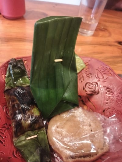 A close-up, eye-level shot shows two Indonesian snacks wrapped in banana leaves, presented on a decorative red plate. The plate has a floral and butterfly pattern. The snacks are situated on a wooden table. To the left, a dark, charred snack, likely grilled, is visible. It is partially wrapped in green banana leaves and secured with a small wooden skewer. To the right, another snack, lighter in color and round, is covered in clear plastic wrap. In the background, a blurred red cup and a partially filled glass of water are present on the wooden table. The scene appears to be in a casual dining setting in Bandarbireuen, Indonesia.