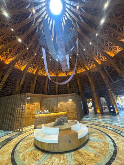 This image captures the interior of a large, high-ceilinged building in Púng Luông, Vietnam, consistent with a resort lobby or public space. The dominant feature is the vaulted, conical ceiling constructed from numerous light-colored wooden or bamboo beams, forming a complex lattice pattern of interconnected triangles and diagonals. Multiple warm-toned lights are integrated into the ceiling structure, illuminating the wooden framework. A large circular skylight is present at the apex of the ceiling, emitting a bright white light. Suspended from the center of the ceiling, below the skylight, is a multi-layered, abstract art installation made of light-colored fabric or material, forming a flowing, sculptural element.
In the foreground, a circular, low-profile seating arrangement with white upholstered cushions surrounds a central feature containing sand and several dark rocks. The floor beneath this seating area is composed of patterned marble or stone, featuring concentric circular and linear designs in shades of golden-brown, green, and dark grey.
Towards the midground, a reception desk made of wood with an illuminated horizontal strip is visible. A female individual, appearing to be staff, stands behind the desk, looking towards the viewer with a slight smile. A computer monitor and a small figurine or plush toy are on the desk. The wall behind the reception desk is textured wood, arranged in block patterns. To the left of the desk, a tall, grid-like wooden partition stands. To the right of the reception area, arched doorways or openings lead to other interior spaces, revealing additional wooden structural elements and distant figures.
