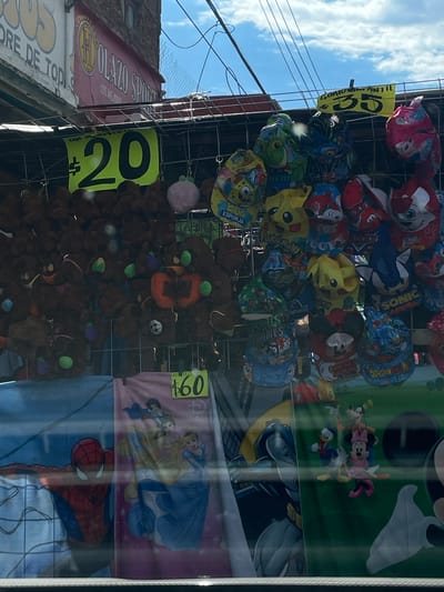 This image shows a storefront display, likely in Oaxaca City, Mexico, featuring a variety of items for sale. The items include character-themed hats featuring designs like Mickey Mouse, Pikachu, and Spiderman, priced at 35 Mexican pesos. There are also plush toys, possibly teddy bears, being sold for 20 Mexican pesos each. Additionally, character-themed towels with designs of Disney princesses and Spiderman are priced at 60 Mexican pesos. The storefront has signage in Spanish. The image appears to be taken from inside a store looking out towards the street, with reflections visible on the glass. A building with the sign "OLAZO SPORT" is visible on the left side of the photo.