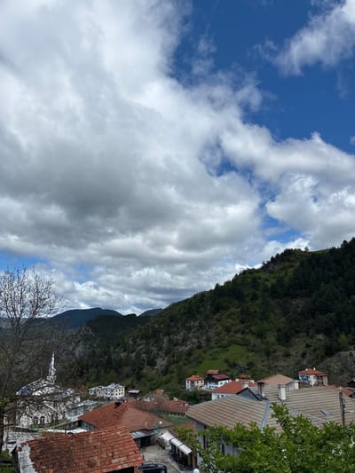 The image shows a view in Giovren, Bulgaria, looking towards a mountainside. The sky is partially cloudy with patches of blue visible. A steep, tree-covered mountainside rises to the right of the frame, with buildings of the town nestled along its base. The buildings have varying roof colors, mostly red and tan. In the foreground are tree branches without leaves.