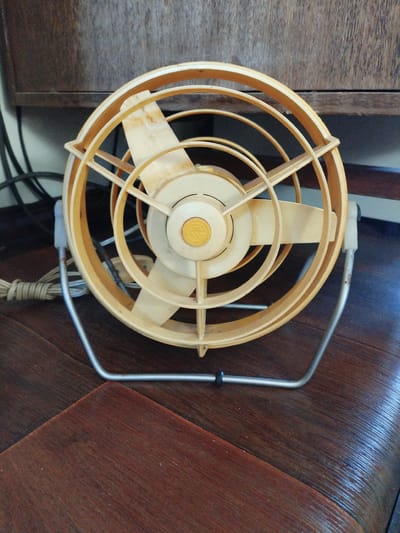A vintage, cream and tan oscillating fan rests on a dark wood surface, likely a table or floor. The fan has a circular grille with concentric rings and a central motor housing. Three fan blades are visible through the grille, and a circular emblem with a stylized "M" is present on the motor. The fan is supported by a silver metal stand. A white power cord with a coiled section is plugged into the fan and trails off to the left. In the background, a dark wooden cabinet with vertical planks is visible. The lighting suggests an indoor setting. The scene is static and shows no people or activity. The location of Вароніцкі сельскі Савет, Belarus is not explicitly depicted or suggested by any elements within the image.