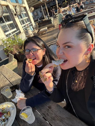 Two smiling women are seated at an outdoor table enjoying food. One woman wears glasses and a dark sweater. The other is wearing a black blazer and lace top with her sunglasses perched on her head. The table is cluttered with empty plastic cups, napkins, and a partially eaten plate of food. A cell phone rests on the table as well. In the background, a building with many windows can be seen.