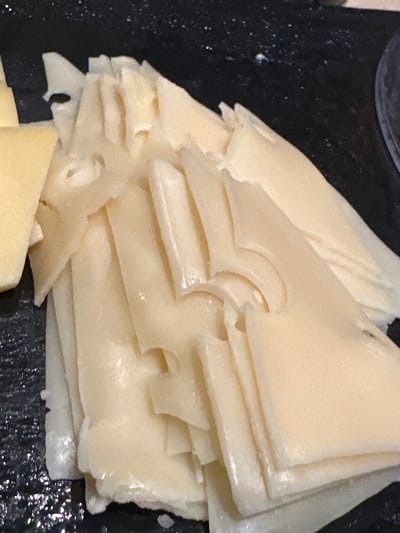 The image presents a close-up, high-angle view of a stack of thinly sliced, light yellow food items, resembling cheese, arranged on a dark, reflective surface. The slices are irregularly shaped, with many exhibiting distinct, rounded perforations or holes, similar to Emmental or Swiss cheese. The texture appears smooth with a slight sheen. A smaller cluster of uniformly shaped, darker yellow slices is visible on the far left. The background consists of a black or very dark platter, showing subtle reflections and sparse, faint white speckles. A dark, circular edge, possibly part of another serving dish, is partially visible on the upper right. No individuals, specific actions, or direct environmental indicators are present. The scene is depicted as taking place in the city of Púng Luông, Vietnam.