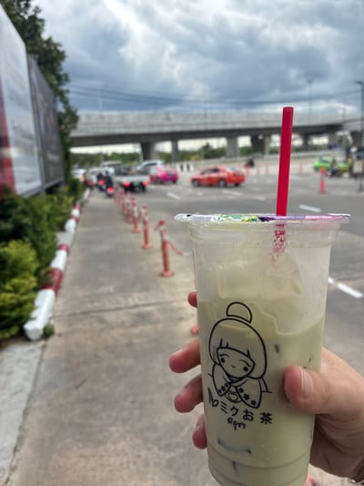 A hand holds a clear plastic cup of a light green beverage, likely iced matcha, with a red straw. The cup has a drawing of a smiling cartoon character with an updo hairstyle, wearing a kimono, and text in an Asian script below it. The cup is positioned in the foreground, obscuring some of the background.
In the background, a road is visible with several vehicles, including a pink car, a red car, a white truck, and motorcycles. There are orange and white traffic cones lining the side of the road and an elevated highway structure above. Lush green trees and foliage are present on the left side of the road, adjacent to a sidewalk or pedestrian path. The sky is overcast with dark clouds.
The scene appears to be a roadside location in the city of Sao Thong Hin Town Municipality, Thailand, possibly near an intersection or along a main thoroughfare given the presence of traffic and the elevated highway. The overall impression is of a casual, possibly urban or suburban, environment during cloudy weather.