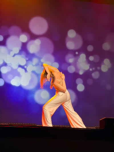 A solo female dancer is captured mid-performance on a stage in the city of Ventspils, Latvia. She has light brown or auburn hair, part of which is styled in a braid visible on her right side, while loose strands frame her face. The dancer is wearing a light beige or off-white, shimmering sleeveless top and matching wide-leg trousers. Distinct blue lines are painted or designed onto her left arm and shoulder. She is positioned in an expressive dance pose, with her back arched, head tilted backward, and arms bent with hands near the back of her head, while maintaining a wide, low stance with her legs.

The stage floor, dark in color, features a horizontal line of recessed or embedded lights along its front edge, emitting a strong orange-red glow that illuminates the dancer's lower body and the immediate stage area. Behind the dancer, a large projection screen displays an atmospheric backdrop. This backdrop consists of a deep blue and purple gradient, overlaid with numerous large, diffused, circular white and light purple lights, creating a bokeh effect. A subtle orange-red tint is visible along the very top edge of the projected image. The scene is dynamically lit, with warm stage lights complementing the cooler, ethereal projection.