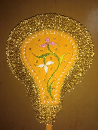 A single, ornate, fan-shaped decorative object is centered against a plain, light brown background. The object features a primary yellow fabric surface adorned with a floral embroidery pattern consisting of a pink flower, a white flower, and green stems/leaves. Small, reflective gold-colored sequins or beads are scattered across the yellow surface, and a border of white dashed stitching outlines the central design area. The entire perimeter of the fan-shaped object is edged with a dense, shimmering gold tinsel fringe. A narrow, cylindrical handle, consistent in color with the main object's backing, extends downwards from the base of the fan. The object's location in Jehonville, Belgium, cannot be determined from the visual evidence within the image.