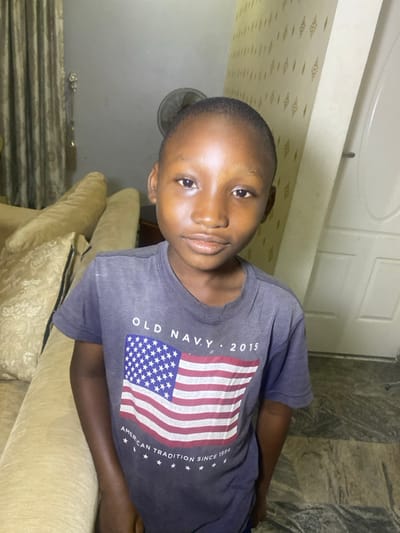 A young boy is resting on a beige sofa. He has dark skin, short dark hair and is wearing a gray t-shirt with an American flag design. The shirt says "Old Navy 2015 American Tradition Since 1994". Behind him are a white door and patterned wallpaper. A fan is partially visible. The lighting is dim.