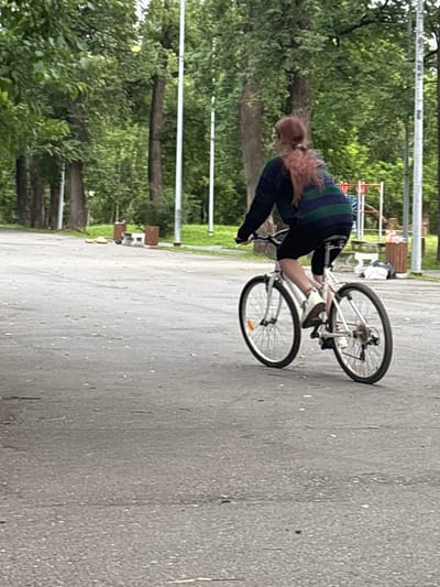 A person with long, light brown hair is riding a white bicycle on an asphalt path. They are wearing a dark blue and green striped shirt and black pants. The bicycle has two wheels with black tires and silver spokes. The background consists of green trees and some park benches. The scene appears to be a park or recreational area, likely outdoors during the daytime. The weather appears to be overcast or cloudy, as the lighting is diffused. The image is taken from a slightly elevated angle, looking down at the person and bicycle.