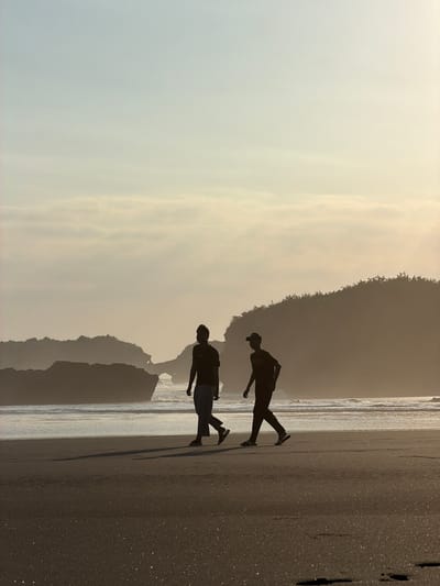 The image displays a coastal scene at a beach in Selemadeg, Indonesia, bathed in the soft, warm glow of either sunrise or sunset, which silhouettes the foreground subjects and distant landforms. Two individuals are walking from left to right across a wide expanse of dark, damp sand in the midground. The person on the left is taller, attired in a t-shirt and loose-fitting pants, and appears to be barefoot or wearing open-toed footwear. The person on the right, slightly shorter, wears a t-shirt, pants, and a baseball cap, with sandals or slippers on their feet. Both individuals cast elongated shadows forward onto the glittering sand.

In the background, the ocean features gentle waves breaking into white foam along the shoreline. Beyond the water, the horizon reveals distinct geological features: to the left, multiple dark, silhouetted rock formations or islands are visible, including a prominent natural rock arch. To the right, a large, dark, undulating hill or coastal mountain, densely covered in vegetation, rises from the ocean's edge. The sky transitions from a light blue at the top to a pale golden yellow near the horizon, with subtle, horizontally stretched cloud formations visible.
