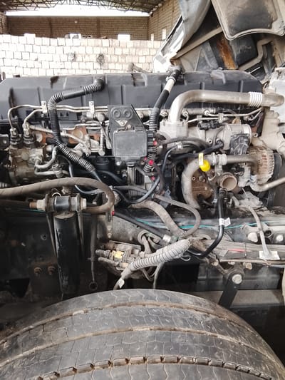 The image shows the engine bay of a truck, likely undergoing maintenance or repair. The engine is a complex array of hoses, wires, and components. A large tire is visible at the bottom of the frame, suggesting the engine is located above the front axle. The setting appears to be a workshop or garage, indicated by the cinder block wall in the background. The image was taken in Sabir Rakhimov, Uzbekistan.