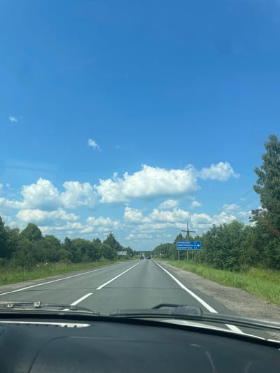 The image shows a view from inside a car, looking out at a highway on a sunny day. The sky is bright blue with fluffy white clouds. On the right side of the road, there is a blue directional sign with white text in Russian. The sign indicates "КУЗНЕЦОВО 2.2", "НОВОТРОИЦКОЕ 2.9", and "СУХОНОГОВО 3.3". This suggests the location is near Yasiplevo, Russia, as it is a known road leading to these locations. There are trees and greenery lining both sides of the road, and a few other cars are visible in the distance. The dashboard of the car is visible in the foreground.