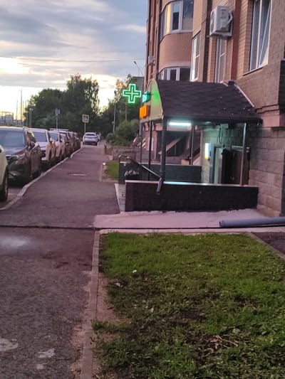 The image shows a street scene in Shchyolkovo, Russia, with a pharmacy sign prominently displayed. The pharmacy features a bright green, illuminated cross sign and orange lettering, indicating its presence. The building itself is a brick structure with multiple windows and an air conditioning unit mounted on the exterior. In front of the pharmacy, there is a covered entrance with artificial lighting. The street is lined with parked cars, mostly SUVs and sedans, on the right side. A sidewalk runs alongside the parked cars, and further to the right, a patch of grass and a curb are visible. The sky above is cloudy with hints of sunset colors. The overall impression is that of a typical urban environment in the late evening.