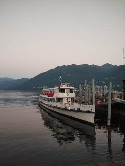 A wide shot captures a ferry named "TORINO" docked in the calm waters of a lake, with a backdrop of dark, tree-covered mountains. The sky is a soft, overcast gray, and the water reflects the ferry and the muted colors of the landscape. The ferry, predominantly white with red accents on its upper deck railings, is positioned parallel to a pier structure. The pier is characterized by several tall, cylindrical grey posts and some docked boats visible in the background to the right. The "TORINO" text is clearly visible on the side of the ferry. The scene appears to be set in Muralto, Switzerland, a location known for its lakefront and surrounding mountains. The image is captured at what appears to be either dawn or dusk, given the soft lighting and the lack of harsh shadows. There are no people visible in the frame.