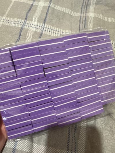 This media file shows a top-down view of a package of purple and white striped nail files. The nail files are arranged in neat rows within a clear plastic wrapper. The background consists of a gray fabric with a plaid pattern, featuring lines in white and dark blue. No people or activities are depicted. The lighting suggests an indoor setting, possibly a home environment. There is no visible text in the image. The context provided indicates the location as Yarominski Village Council, Belarus.