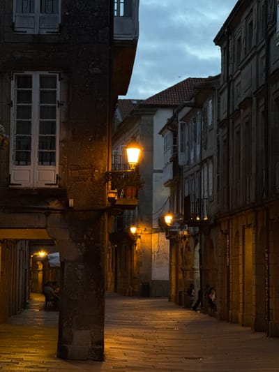 This image depicts a narrow street in Santiago de Compostela, Spain, during dusk or early evening. The architecture suggests an old European city, with weathered stone buildings lining the street. The sky, visible between the buildings, is cloudy, indicating overcast weather.
The scene is illuminated by warm, artificial lights, including street lamps and lights emanating from within the buildings. Several people are visible in the distance, walking or standing, adding a sense of life to the otherwise somber atmosphere. One person appears to be sitting or resting on a bench. The mood is quiet and contemplative, perhaps due to the time of day and the enclosed nature of the street.
Notable details include the texture of the stone buildings, the tiled roofs, and the windows on the facades. The perspective of the photograph is somewhat unusual, as if taken from a low angle or with a tilt. The image captures the architectural character of the historic city and the atmospheric quality of its streets after sunset.
