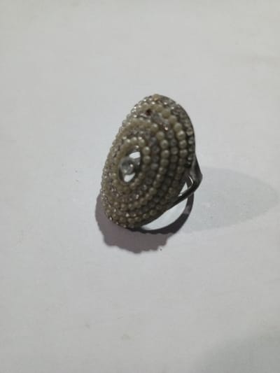 The image displays a single, elongated, oval-headed ring positioned on a flat, light-colored surface, reportedly in the city of Phakeo, Laos. The ring's head is densely encrusted with multiple concentric rows of small, round, cream-colored beads or simulated pearls. A single, larger, clear, faceted gemstone is centrally set within the beaded design. The metallic band of the ring is visible, resting on the surface and casting a distinct shadow to the lower-right, indicating illumination from the upper-left. The background surface is uniformly light, exhibiting minor irregularities and dust specks. No individuals or other objects are present in the scene.