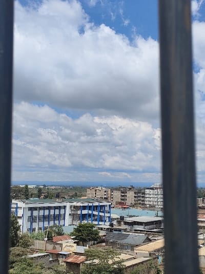 The image presents an elevated, wide-angle view of an urban landscape within Juja ward, Kenya, framed by two prominent, out-of-focus vertical metallic bars on the left and right edges. The upper two-thirds of the scene depict a partly cloudy blue sky, featuring large, white cumulus clouds. Below the sky, the horizon reveals a distant, hazy landmass, possibly a mountain range or hills.

The midground is dominated by a dense arrangement of buildings. A distinctive multi-story building with a white exterior and vertical blue architectural accents is visible on the mid-left. To its right, and extending into the background, are numerous other multi-story structures, several of which appear to be under construction, characterized by exposed concrete frames and rebar. The lower midground consists of closely packed smaller residential and commercial buildings, many featuring corrugated iron roofs, some with visible signs of rust, and others with flat roofs often supporting water tanks. Interspersed throughout the built environment are various green trees and patches of foliage. No individuals or specific activities are discernible within the frame.